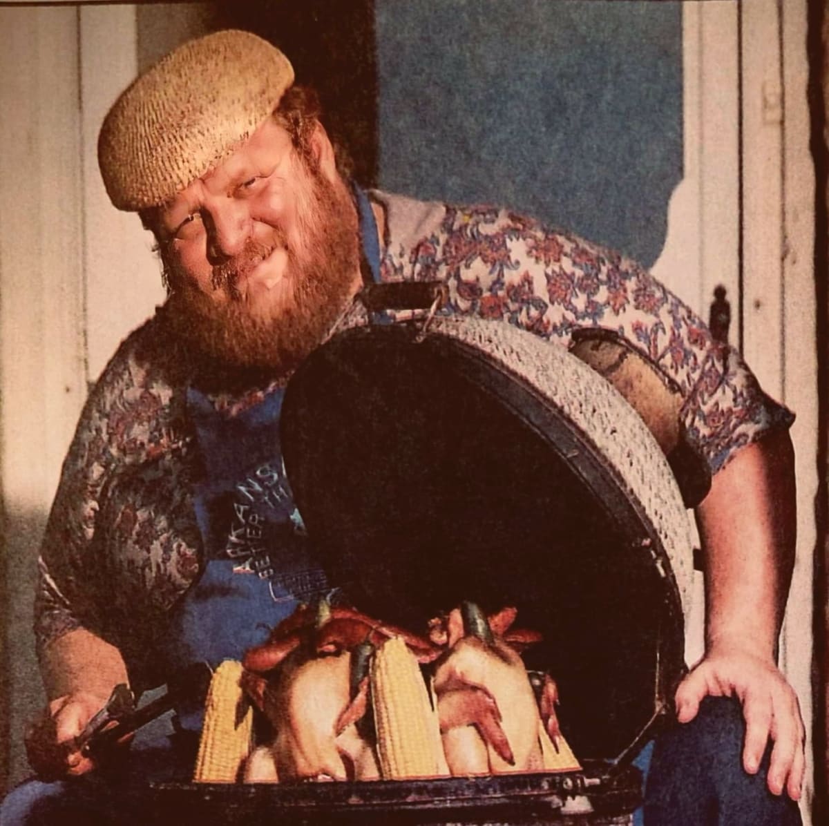 A man in a blue apron holds a black grill full of delicious food on corn on the cob.